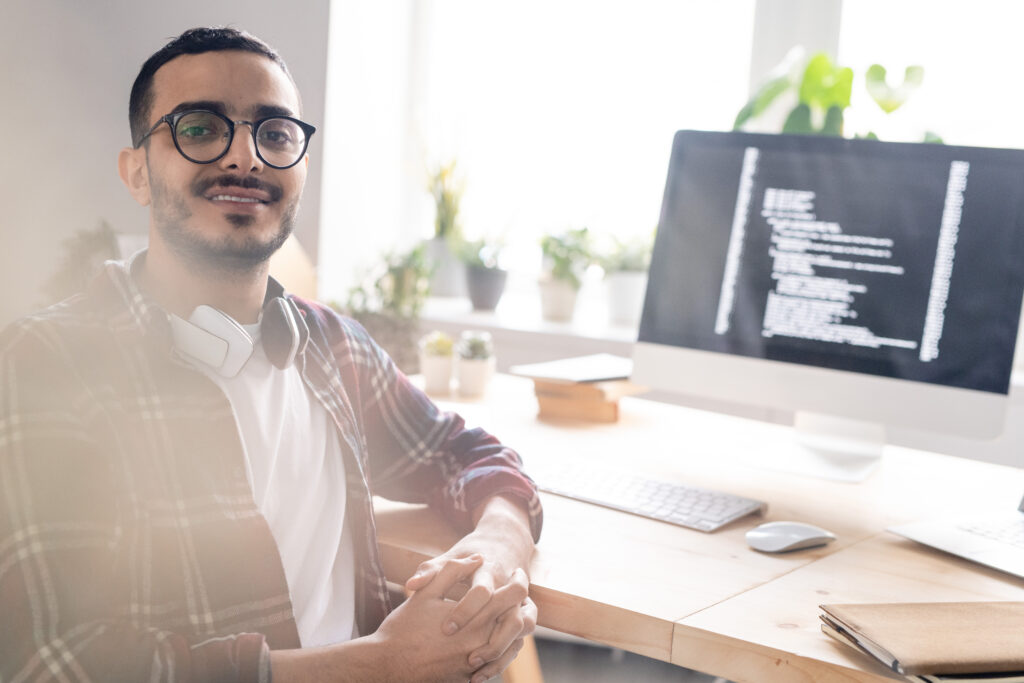 Retrato de jovem programador positivo do Oriente Médio em óculos sentado com as mãos entrelaçadas na mesa com um computador poderoso